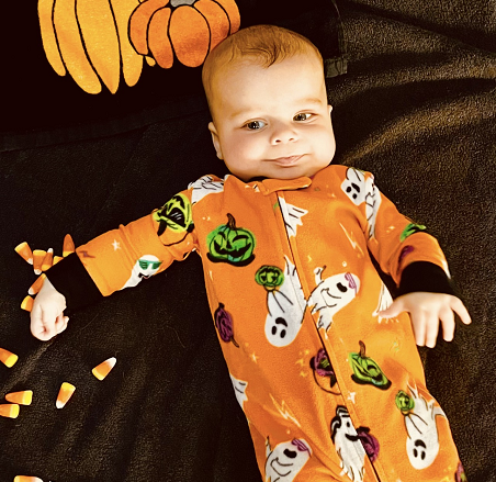 A cute baby dressed in Halloween pajamas laying down on a staged sheet with various Halloween accoutrement