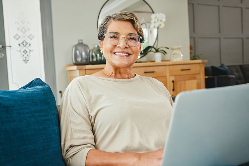 An aged woman looks up from her computer and smiles. She's in a cozy, clean living room.