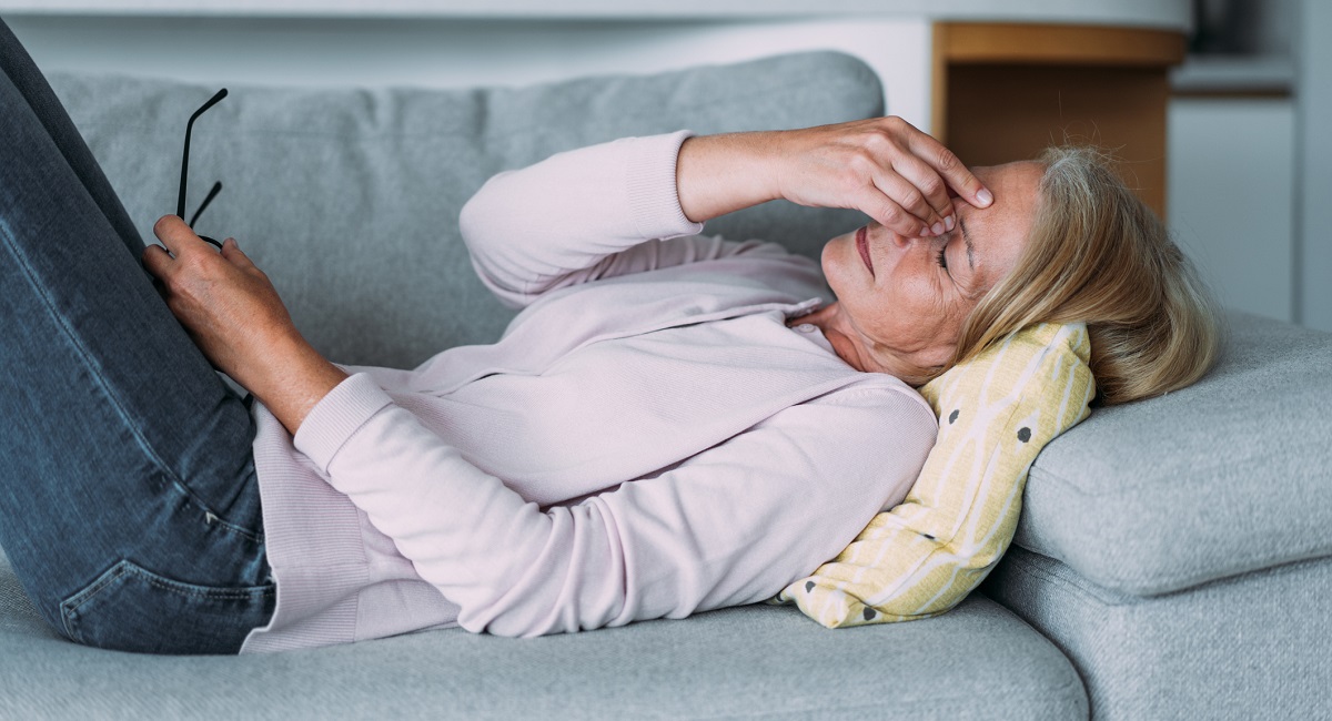 Mature woman laying on couch with a headache and holding the bridge of her nose to ease the pain.