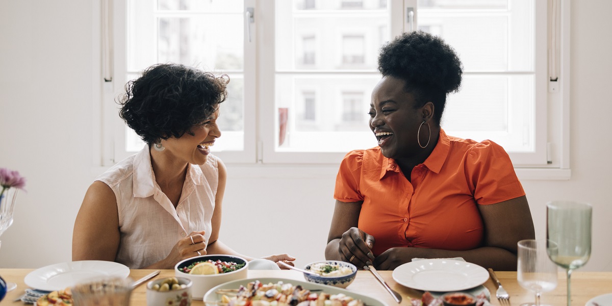 Two women laughing and having lively conversation while sharing a meal in a brightly lit room next to a window during the day