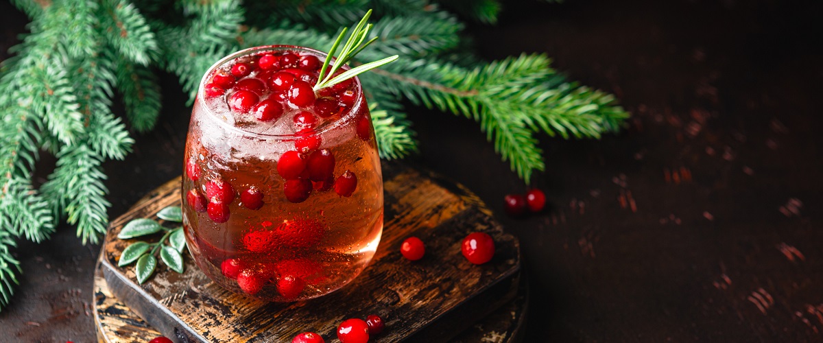 Cranberry mocktail with ice on top of dark wood pieces surrounded by white pine branches and cranberries.