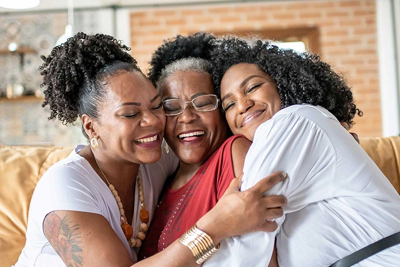 Three women embrace joyfully on a couch, touching their heads together while smiling and laughing